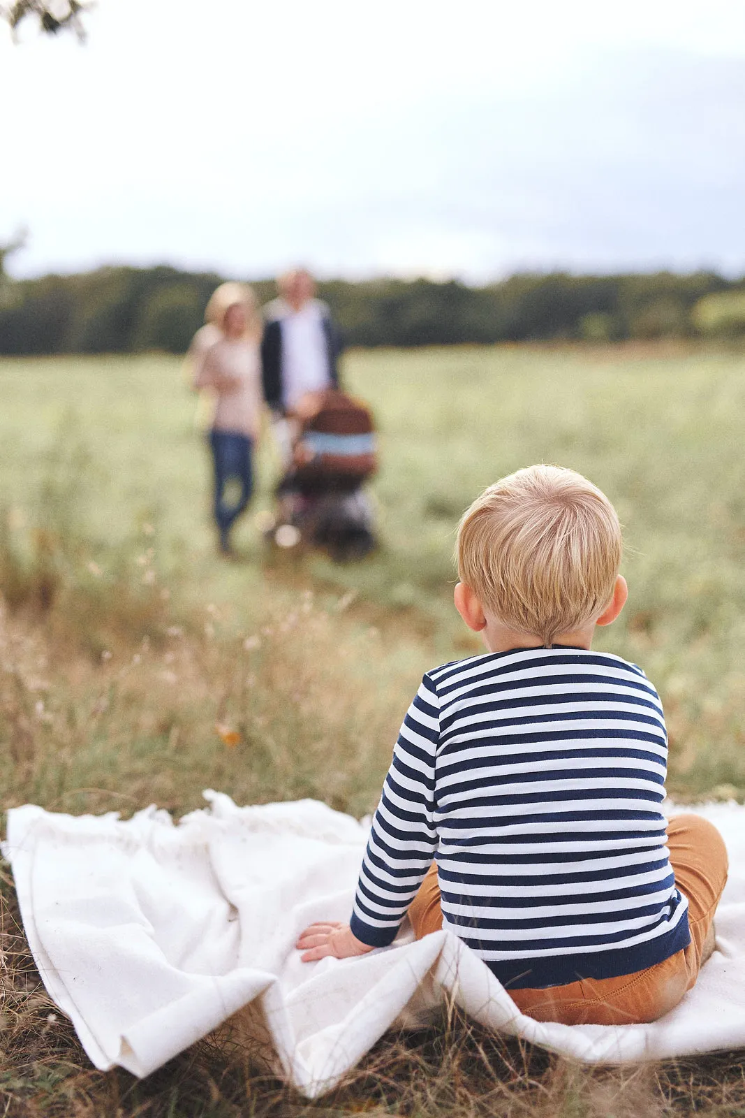 Im Vordergrund ist ein Junge zu sehen der auf einer Decke sitzt und zu seinen Eltern schaut, welche im Hintergrund einen Kinderwaagen schieben.
