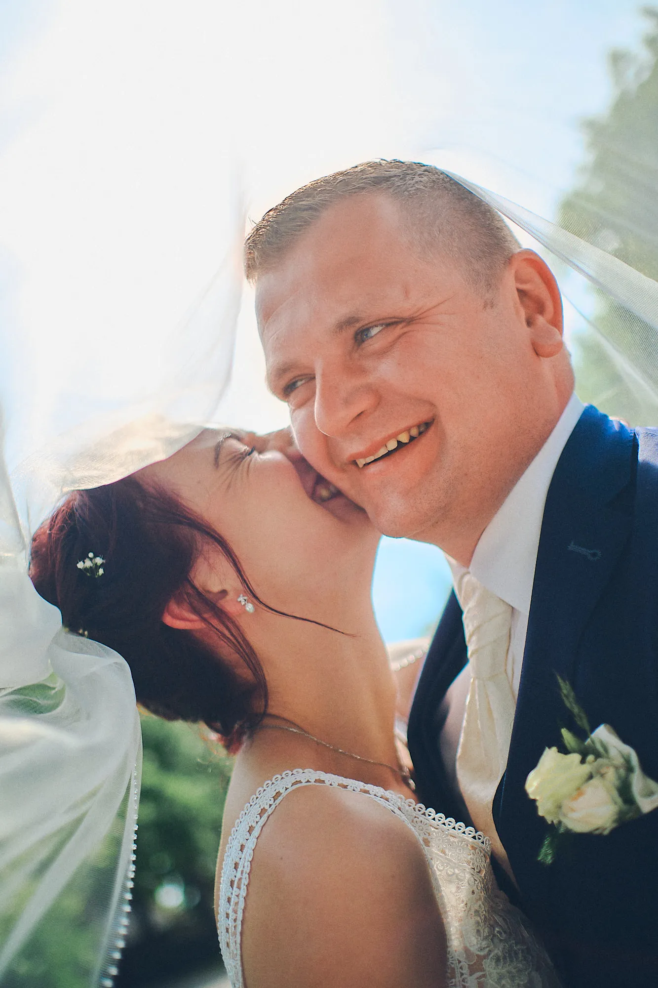 Ein Hochzeitspaar unter einem lichtdurchfluteten Schleier. Die Braut küsst den Bräutigam der sich sehr darüber freut.