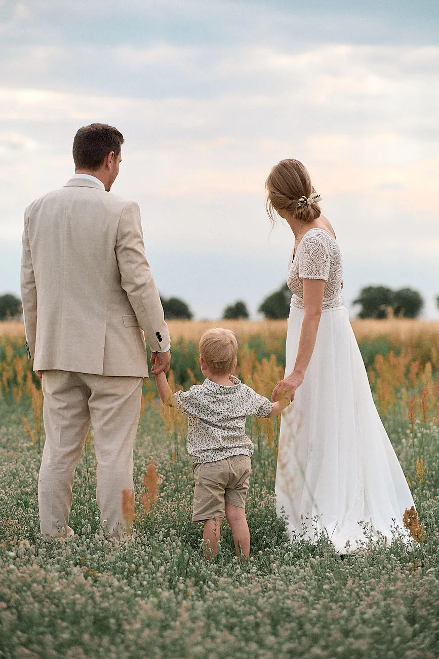 Ein Hochzeitspaar steht auf  einer blühenden Wiese, schaut gemeinsam zum Dämmerungslicht und hält dabei ihr gemeinsames Kind in der Mitte.